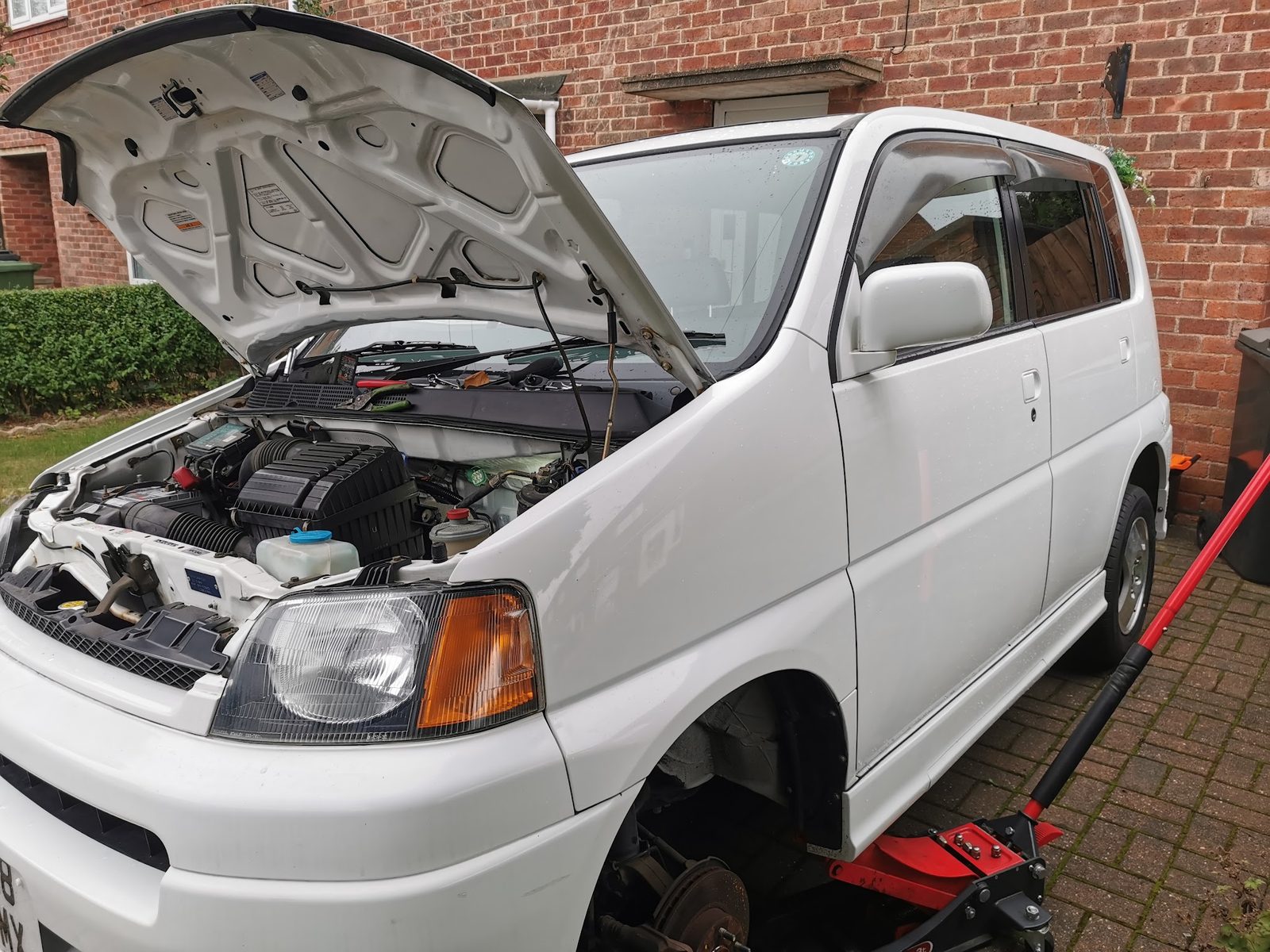 White people carrier on a home driveway with the bonnet up and a front wheel off, mid repair